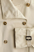 Beige short trench coat for women, featuring a belted waist, buttoned front, and lightweight fabric for versatile styling.
