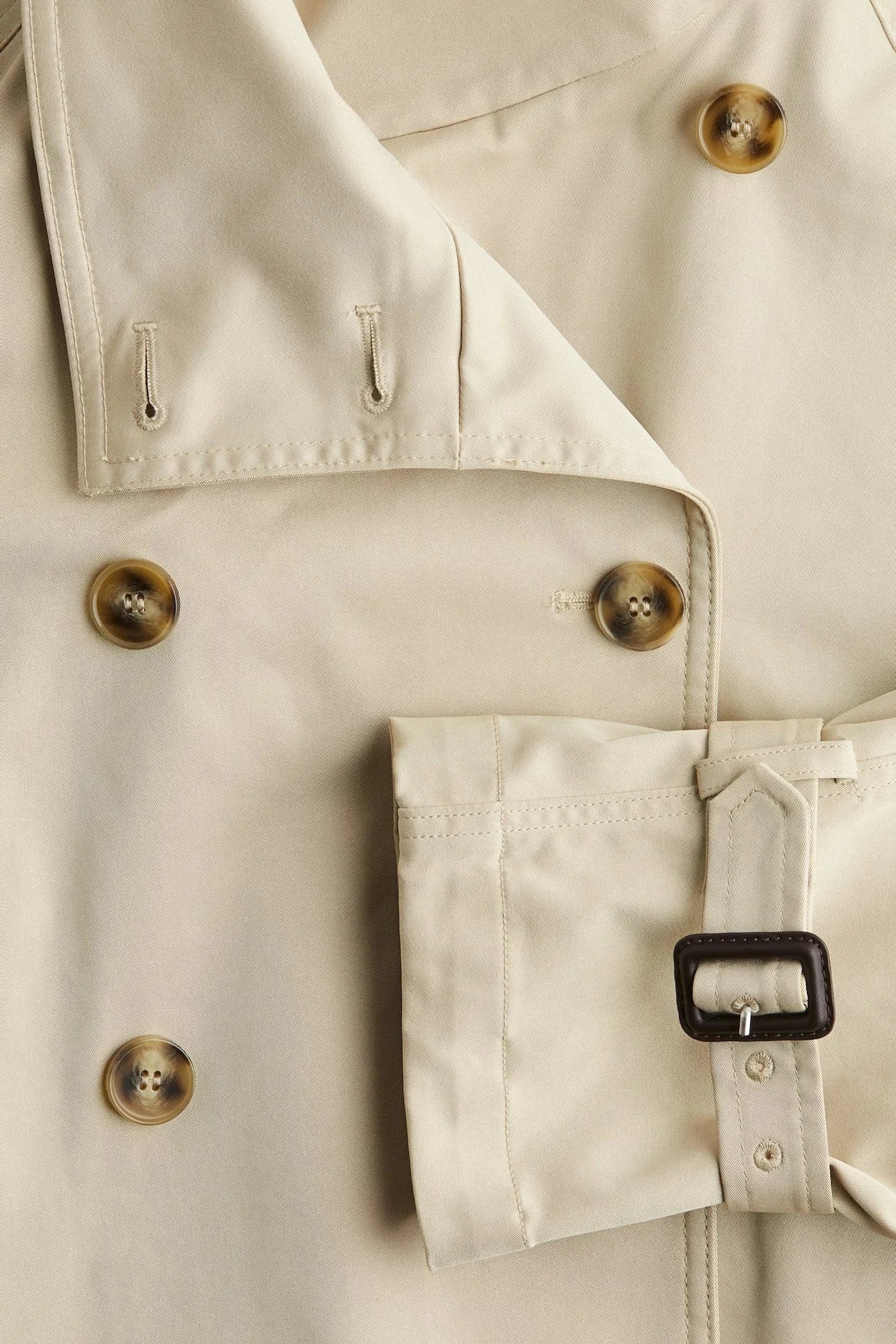 Beige short trench coat for women, featuring a belted waist, buttoned front, and lightweight fabric for versatile styling.
