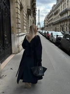 Women's long black coat made of durable fabric, featuring a belted waist and wide lapels for a stylish silhouette.