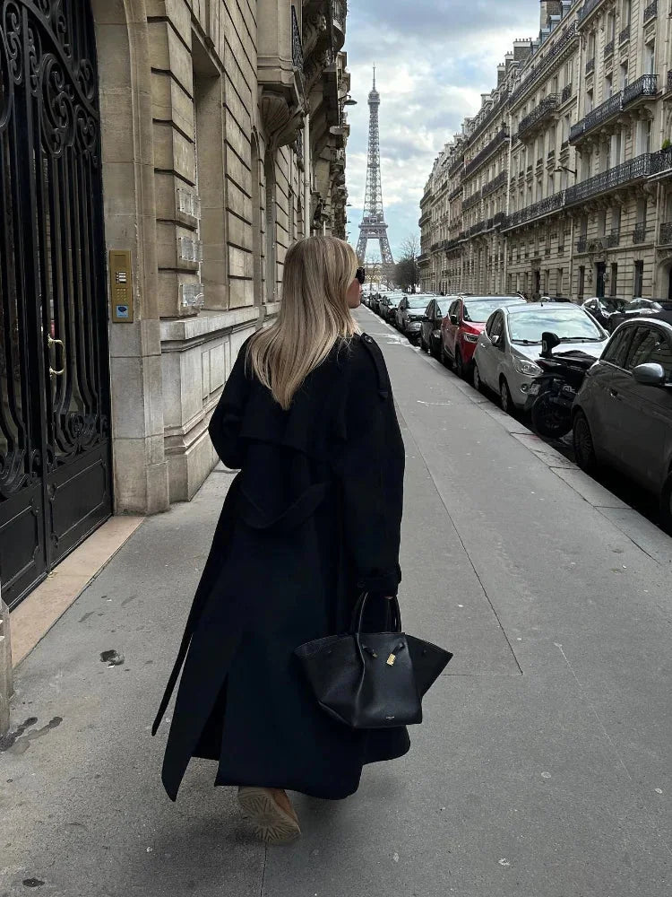 Women's long black coat made of durable fabric, featuring a belted waist and wide lapels for a stylish silhouette.